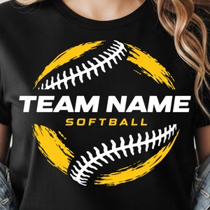 Peut inclure: T-shirt noir avec un motif de softball. Le motif présente un graphisme de softball jaune et blanc avec les mots "TEAM NAME" au-dessus de "SOFTBALL" en lettres blanches.