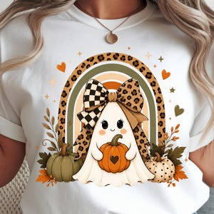 Peut inclure: Un t-shirt blanc avec un motif représentant un fantôme portant un grand nœud à carreaux et imprimé léopard. Le fantôme tient une citrouille orange avec un cœur découpé, devant un arc-en-ciel et entouré de feuillages aux couleurs automnales.