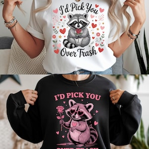 May include: White t-shirt with a raccoon graphic holding a rose, surrounded by hearts and flowers, and the text "I'd Pick You Over Trash." A black sweatshirt features a similar design with a pink raccoon wearing heart-shaped glasses.