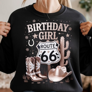 Puede incluir: Sudadera negra con un diseño gráfico que presenta el texto "BIRTHDAY GIRL" encima de un letrero de la Ruta 66. El diseño incluye botas de vaquero, un sombrero, un cactus y estrellas. El diseño es en tonos marrones y blancos.