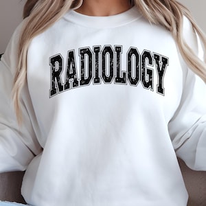 Peut inclure: Sweat-shirt blanc avec le mot "RADIOLOGY" imprimé en noir sur le devant, dans une police de style universitaire vieillie. Le sweat-shirt a un col rond et des manches longues. Le style général est décontracté.