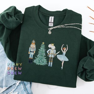 Puede incluir: Una sudadera verde oscuro con un diseño de acuarela de personajes del ballet El Cascanueces. El diseño incluye un cascanueces, un rey ratón, un árbol de Navidad y una bailarina. El texto "FINNY DREW CREW" está impreso en colores en la parte inferior izquierda.