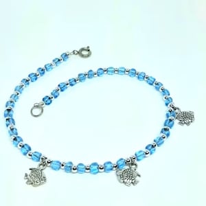 May include: Anklet featuring light blue, faceted beads interspersed with small silver beads. The anklet has three silver fish charms and a silver clasp. The beads are strung on a silver chain, creating a delicate and stylish accessory.