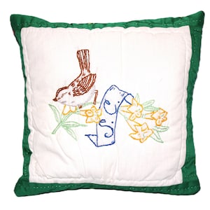 May include: Square white pillow with a green border. The pillow features an embroidered design of a brown bird, a blue banner with text, and yellow flowers with green stems. The pillow is quilted.