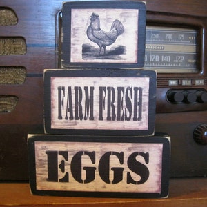 May include: Three wooden signs stacked on top of each other. The top sign has a black and white illustration of a rooster. The middle sign says "FARM FRESH" in black letters on a white background. The bottom sign says "EGGS" in black letters on a white background.