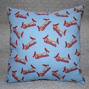 Puede incluir: Una almohada azul claro con un patrón repetido de cardenales rojos posados ​​en bates de béisbol amarillos. La palabra "Cardinals" está impresa en blanco en cada bate.