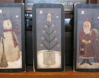 Puede incluir: Tres placas de madera con marcos negros y escenas pintadas. La primera placa presenta un muñeco de nieve con una bufanda roja y una escoba. La segunda placa presenta un árbol de Navidad en maceta con adornos blancos. La tercera placa presenta a un Papá Noel vestido de rojo sosteniendo una linterna y un manojo de palos.