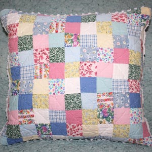 May include: A square throw pillow featuring a patchwork design composed of numerous small fabric squares in various pastel colors and floral, plaid, and solid patterns. The pillow has a white pom-pom trim along the edges and visible quilting stitches.