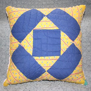 May include: Square pillow with a quilted design. The pillow features a yellow background with a floral pattern and blue quilted shapes. The edges are trimmed with white fabric.