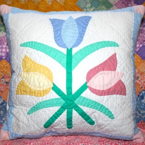 May include: Square quilted pillow with a white background and a floral design. The design features a blue tulip, a yellow tulip, and a pink tulip with green stems and leaves. The pillow has a light blue border.