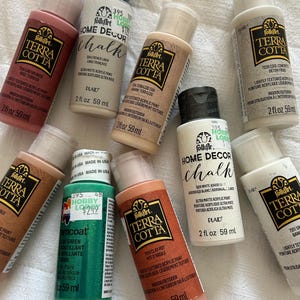 May include: A collection of FolkArt Terra Cotta and Home Decor Chalk acrylic paints in various colors, including terracotta, tan, and green. The bottles are 2 fl oz (59 ml) each and are labeled for indoor/outdoor use. The image shows the bottles on a white surface.