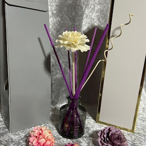 May include: A diffuser bottle with purple reeds and a white flower on top. The bottle is dark grey and has a heart-shaped design. There are three small pink and purple flowers on the table.