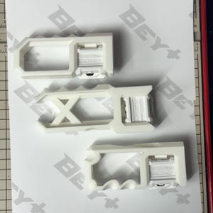 May include: Three white plastic Beyblade X launcher grips with distinct geometric cutouts and ergonomic handle designs. Each grip features a rectangular attachment point with the embossed text BEYBLADE X, arranged vertically on a white surface.