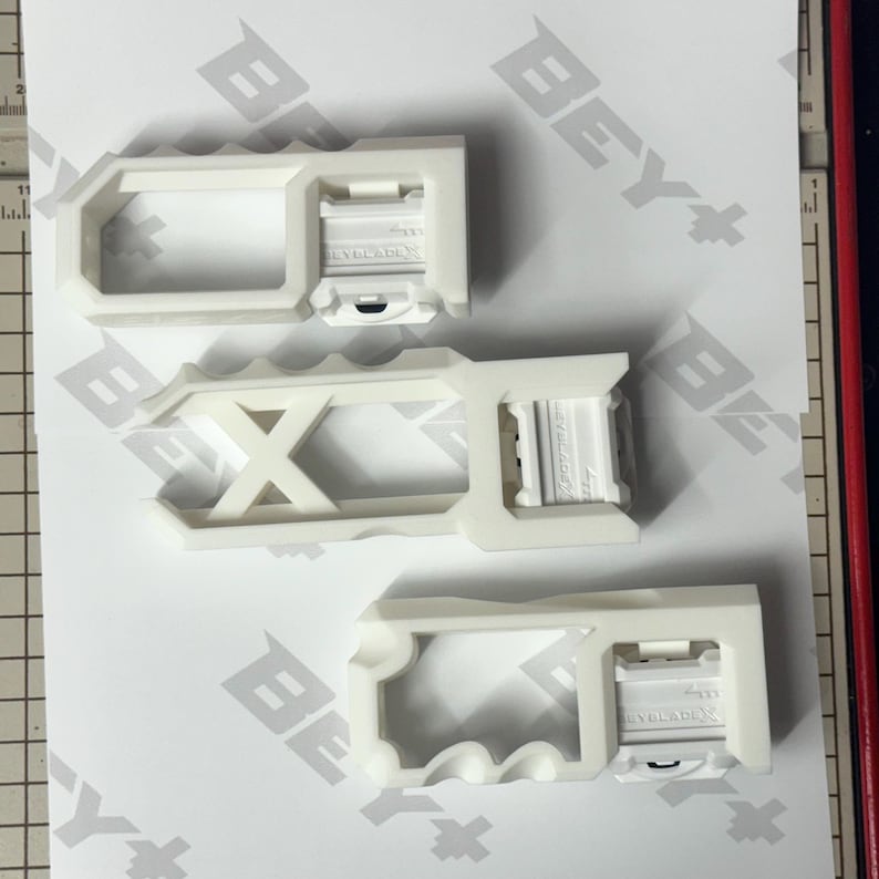 May include: Three white plastic Beyblade X launcher grips with distinct geometric cutouts and ergonomic handle designs. Each grip features a rectangular attachment point with the embossed text BEYBLADE X, arranged vertically on a white surface.