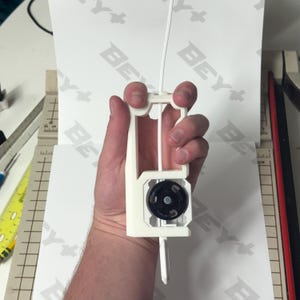 May include: A white 3D-printed plastic filament guide tool featuring a central black circular wheel mechanism. The device has an elongated rectangular frame with a vertical slot for filament to pass through, designed to assist with 3D printer setups.