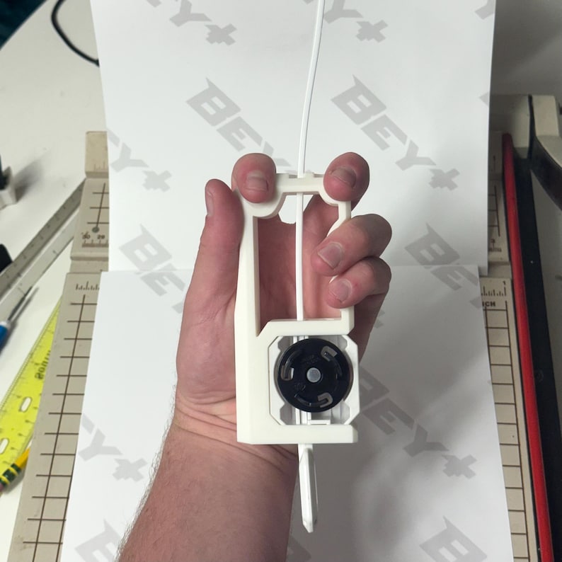 May include: A white 3D-printed plastic filament guide tool featuring a central black circular wheel mechanism. The device has an elongated rectangular frame with a vertical slot for filament to pass through, designed to assist with 3D printer setups.