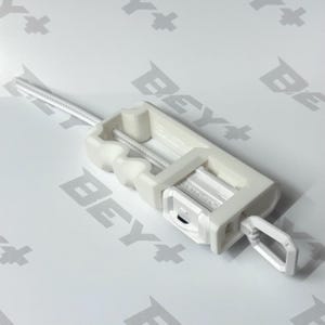 May include: A white plastic Beyblade ripcord launcher with a long, thin, serrated pull strip extending from the top. The rectangular device features a finger ring at the base and the word BEYBLADE embossed on the side of the central mechanism.