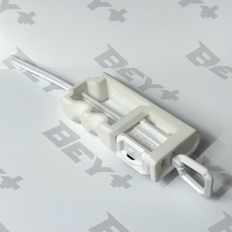 May include: A white plastic Beyblade ripcord launcher with a long, thin, serrated pull strip extending from the top. The rectangular device features a finger ring at the base and the word BEYBLADE embossed on the side of the central mechanism.