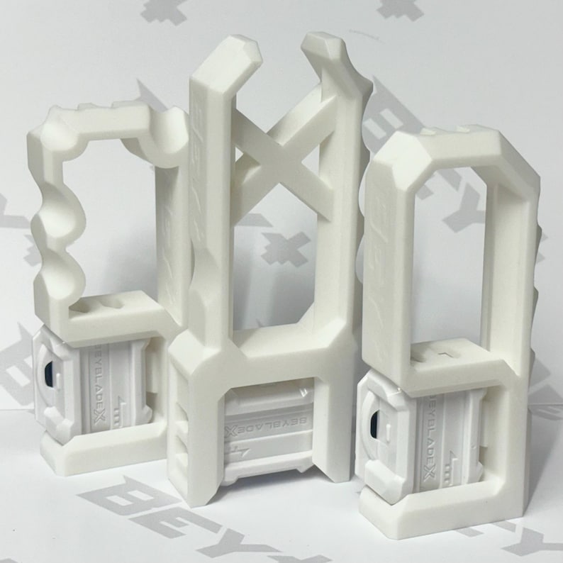 May include: Three white plastic Beyblade X string launcher grips with ergonomic handles and geometric cutouts. Each grip features a central rectangular housing with the embossed text BEYBLADE X, designed for attachment to a compatible launcher.