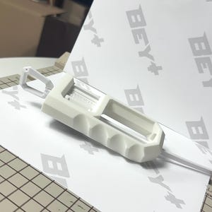 May include: A white 3D printed plastic tool featuring a rectangular body with a series of finger-grip indentations along one side and a small triangular loop at the top. The object is positioned on a white surface with the repeating text BEY+.