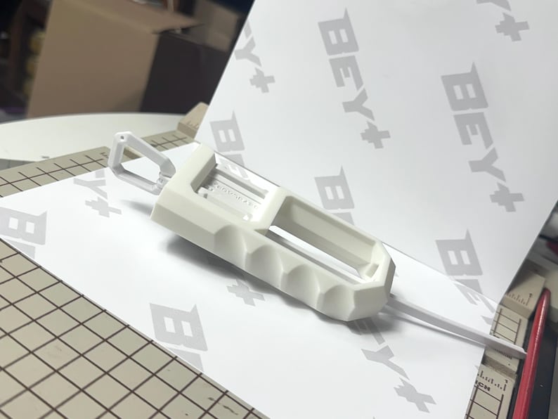 May include: A white 3D printed plastic tool featuring a rectangular body with a series of finger-grip indentations along one side and a small triangular loop at the top. The object is positioned on a white surface with the repeating text BEY+.