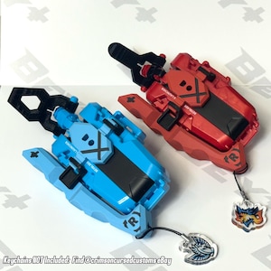 May include: Two toy launchers, one red and one blue, with black accents. Each launcher has a handle and a design with the word "Beyblade" and an "X" symbol. The blue launcher has a small keychain attached. The text "Keychains NOT Included" is at the bottom.