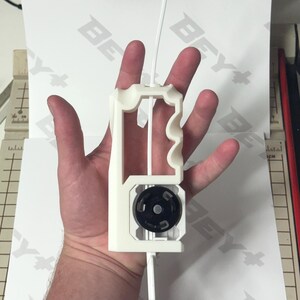 May include: A white 3D-printed plastic launcher tool featuring a vertical handle with ergonomic finger grooves and a circular black gear mechanism at the base. A thin white plastic strip is inserted through the center of the device.