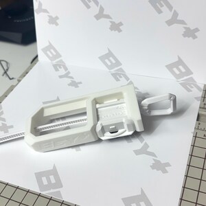 May include: A white 3D printed Beyblade launcher accessory featuring a rectangular frame with the text BEY+ embossed on the side. The device includes a sliding mechanism and a small loop attachment, positioned on a white surface with BEY+ branding.