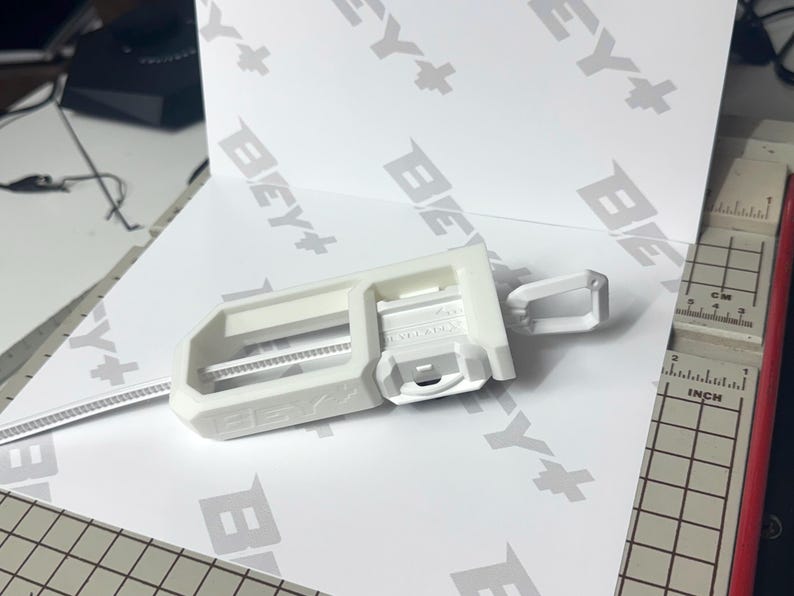 May include: A white 3D printed Beyblade launcher accessory featuring a rectangular frame with the text BEY+ embossed on the side. The device includes a sliding mechanism and a small loop attachment, positioned on a white surface with BEY+ branding.