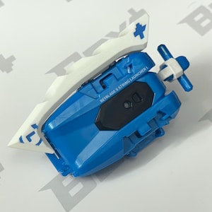 May include: A blue and white Beyblade X String Launcher L toy. The launcher has a blue body with a black accent and white details, including a cross symbol. The text "BEYBLADE X STRING LAUNCHER L" is printed on the side.