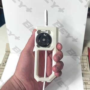 May include: A white 3D-printed plastic launcher tool featuring a central black circular gear mechanism. The device has a rectangular handle frame with a vertical slot for a white plastic strip. The tool is held upright against a white background.