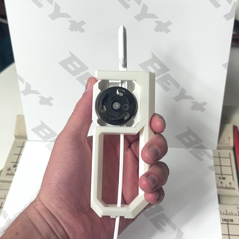 May include: A white 3D-printed plastic launcher tool featuring a central black circular gear mechanism. The device has a rectangular handle frame with a vertical slot for a white plastic strip. The tool is held upright against a white background.