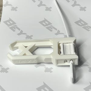 May include: A white plastic Beyblade launcher tool featuring an elongated rectangular frame with an X-shaped support structure and a side-mounted pull cord mechanism. The device is embossed with the text BEY+ on the side and Beyblade on the top.