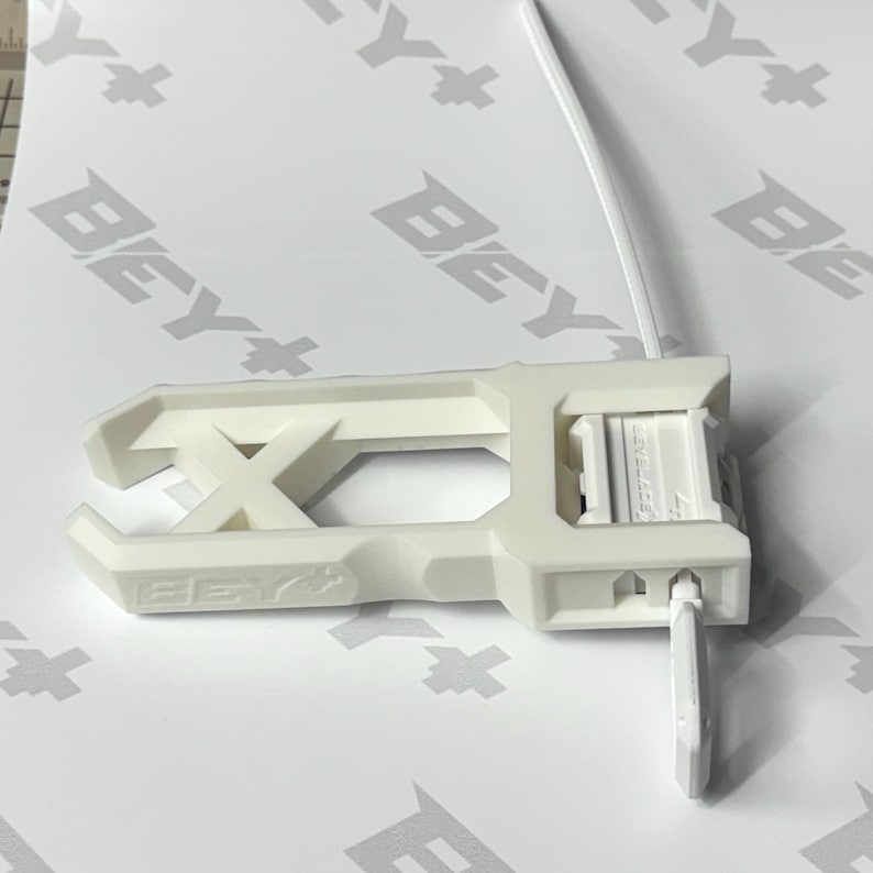 May include: A white plastic Beyblade launcher tool featuring an elongated rectangular frame with an X-shaped support structure and a side-mounted pull cord mechanism. The device is embossed with the text BEY+ on the side and Beyblade on the top.