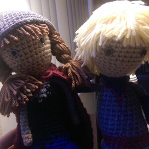 May include: Two crocheted dolls, one with brown hair and a purple hat, the other with blonde hair. Both dolls are wearing colorful clothing.