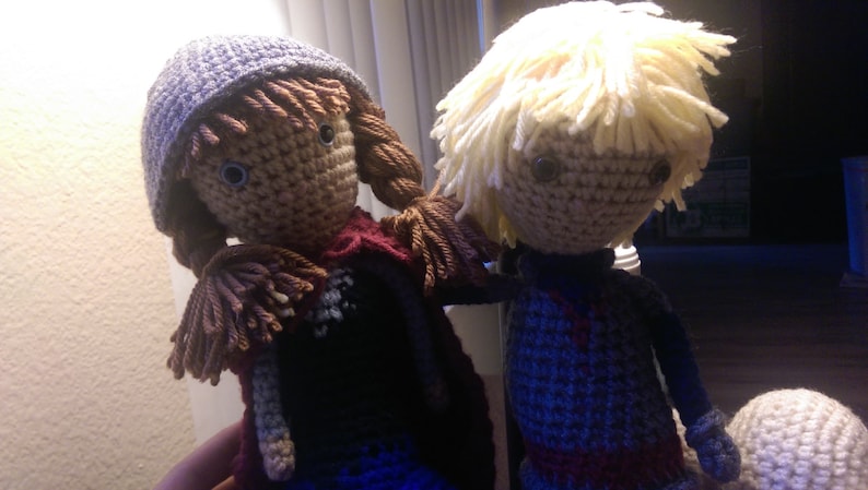 May include: Two crocheted dolls, one with brown hair and a purple hat, the other with blonde hair. Both dolls are wearing colorful clothing.