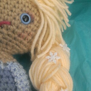 May include: A crocheted doll with long blonde hair styled in a braid. The doll is wearing a blue crocheted dress and has white snowflake embellishments in her hair.