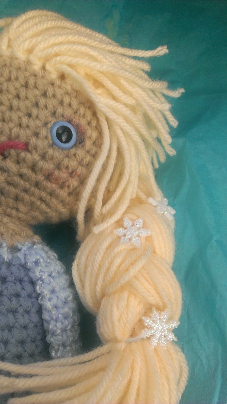 May include: A crocheted doll with long blonde hair styled in a braid. The doll is wearing a blue crocheted dress and has white snowflake embellishments in her hair.