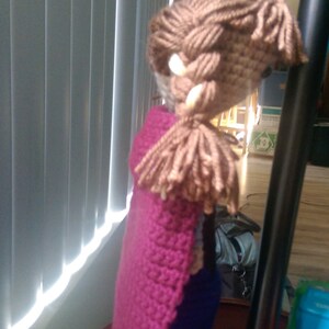 May include: A crocheted doll with brown hair, a pink cape, and a blue dress.