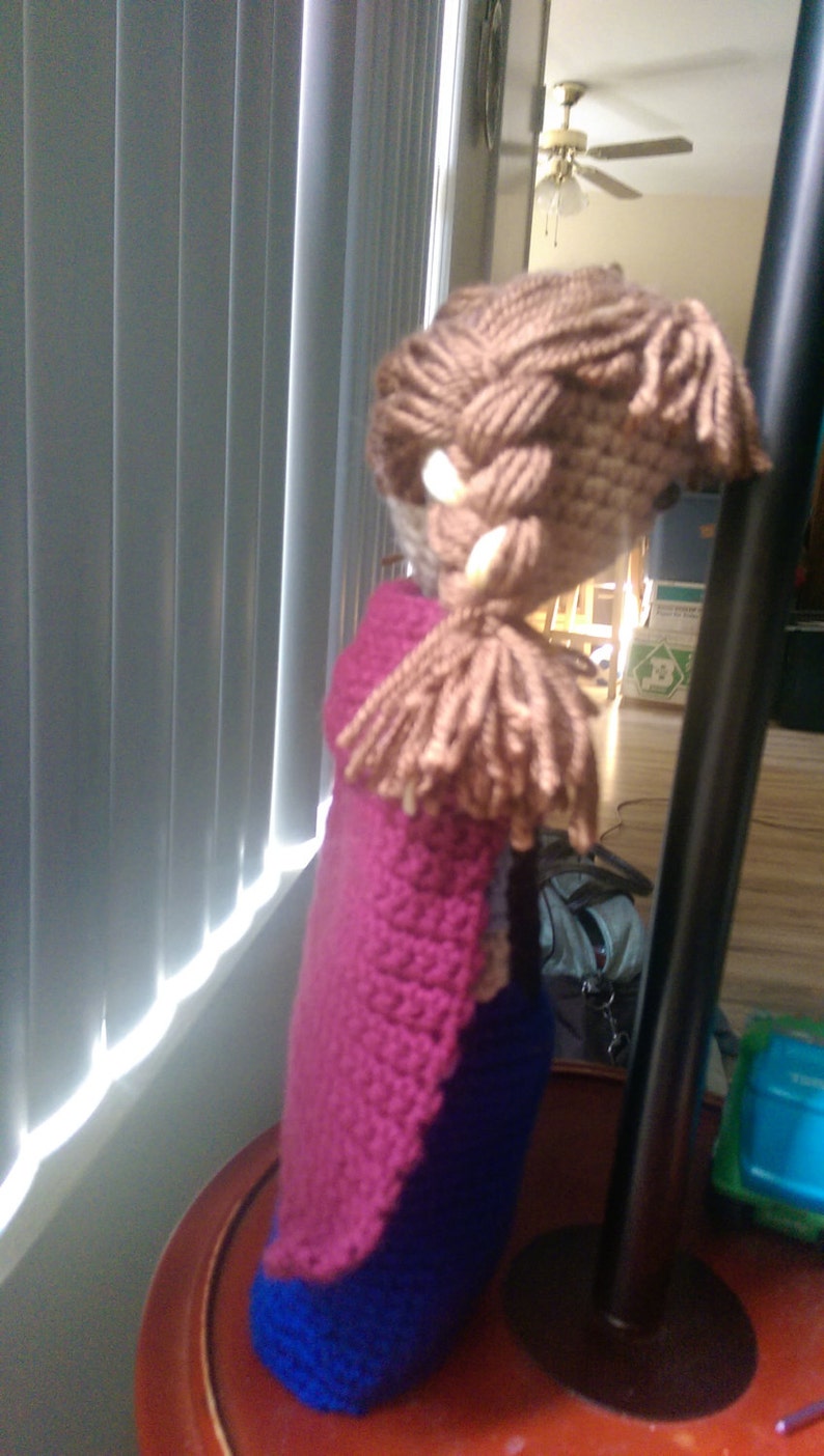 May include: A crocheted doll with brown hair, a pink cape, and a blue dress.