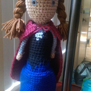 May include: Crocheted doll with brown hair, blue eyes, and a red cape. The doll is wearing a blue dress and is standing on a red surface.