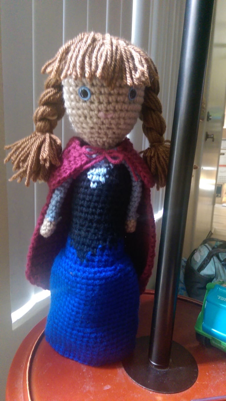 May include: Crocheted doll with brown hair, blue eyes, and a red cape. The doll is wearing a blue dress and is standing on a red surface.