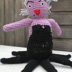 Könnte beinhalten: Handgefertigte Häkelpuppe, die eine Figur mit violetter Haut, einem schwarzen Kleid und grauem Haar darstellt. Die Puppe hat große weiße Augen, eine rote dreieckige Nase und schwarze Tentakel. Ein einzigartiger, handgefertigter Artikel.