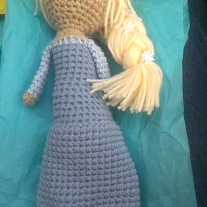 May include: A crocheted doll with long blonde hair, wearing a light blue dress. The doll has blue eyes and a pink mouth. The doll is sitting on a light blue tissue paper.