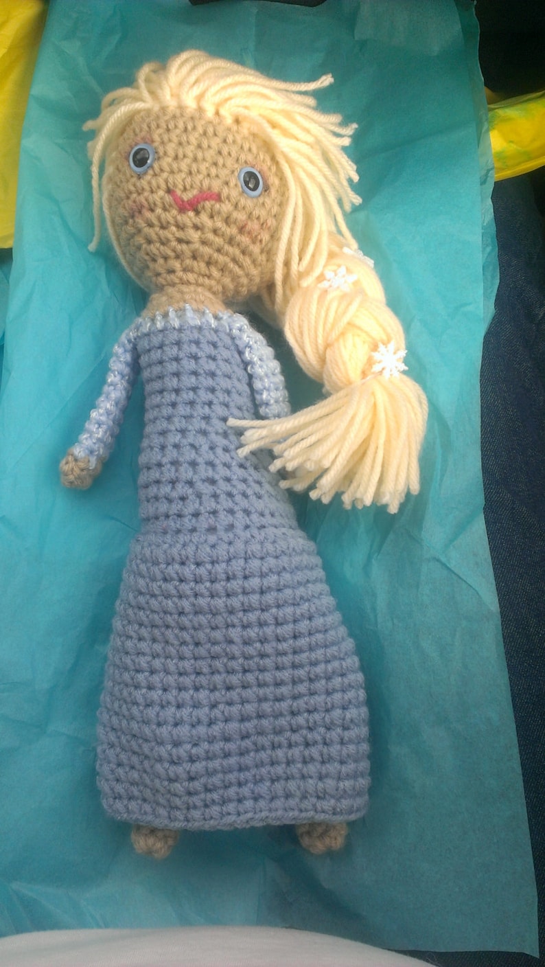 May include: A crocheted doll with long blonde hair, wearing a light blue dress. The doll has blue eyes and a pink mouth. The doll is sitting on a light blue tissue paper.