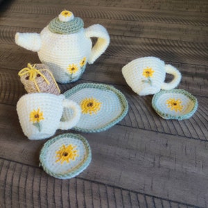 Könnte beinhalten: Ein gehäkeltes Teeservice in Creme und Gelb, bestehend aus einer Teekanne, Tassen, Untertassen und einem kleinen rechteckigen Gegenstand. Jedes Teil ist mit einem gelben Sonnenblumenmuster verziert. Das Set ist auf einer Holzoberfläche angeordnet.