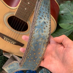 May include: A blue guitar strap with gold star accents and a brown and white herringbone border. The strap is held in front of a wooden acoustic guitar.