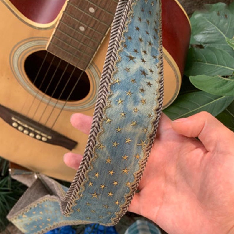 May include: A blue guitar strap with gold star accents and a brown and white herringbone border. The strap is held in front of a wooden acoustic guitar.