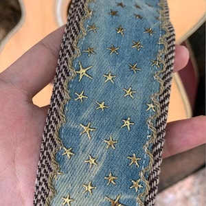 May include: A denim guitar strap with gold star embroidery. The strap has a blue denim center with gold star accents and a brown and tan woven border. The strap is held in a hand.