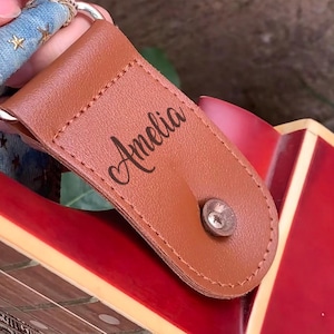 May include: A brown leather guitar pick holder with the name "Amelia" printed on it. The holder is attached to a blue and white patterned strap with a silver metal ring. The guitar body is red.